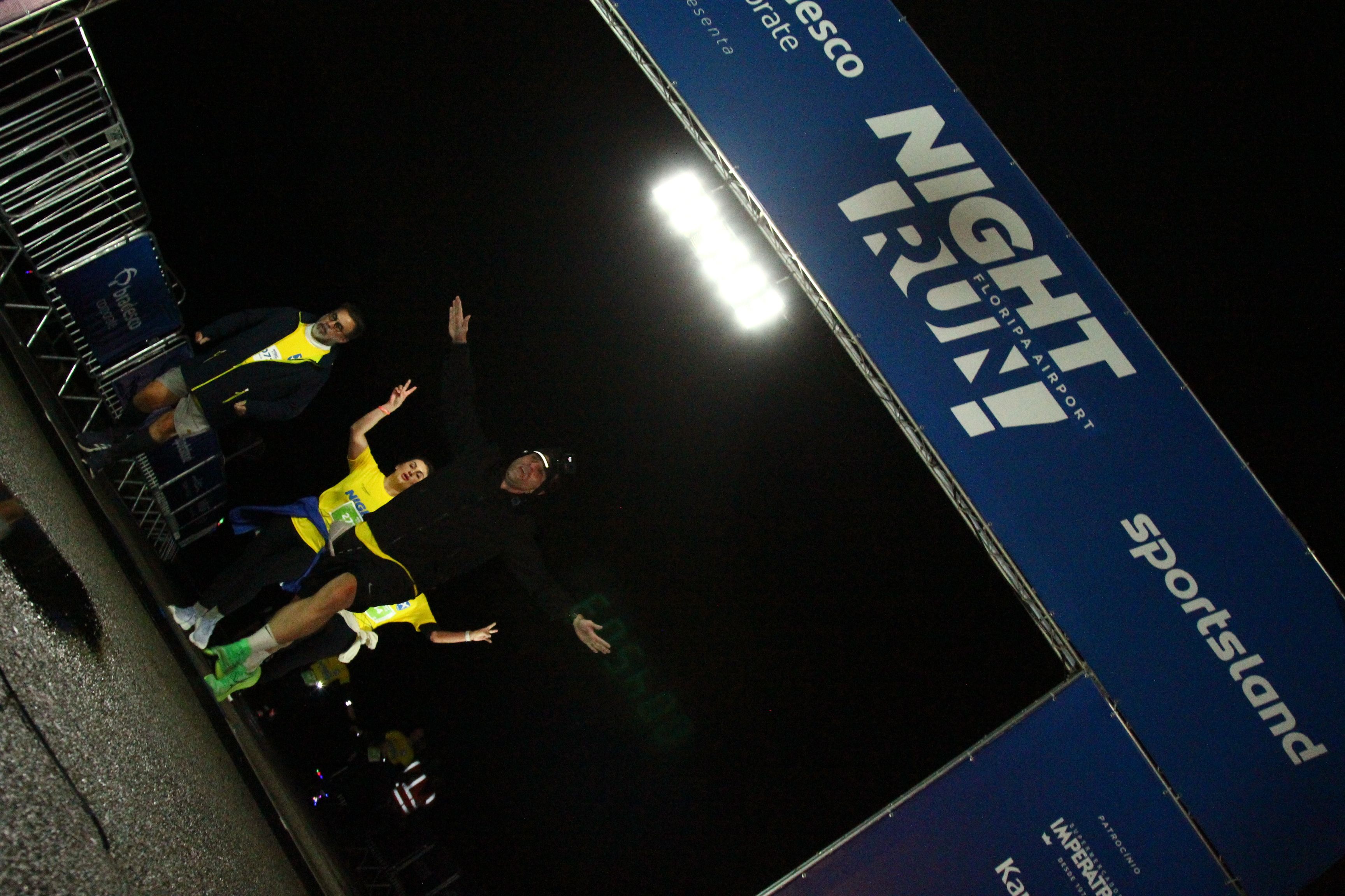 Arco de largada da Night Run Floripa Airport iluminado à noite, com o letreiro NIGHT RUN Floripa Airport e a marca Sportsland, chão molhado de chuva refletindo a luz