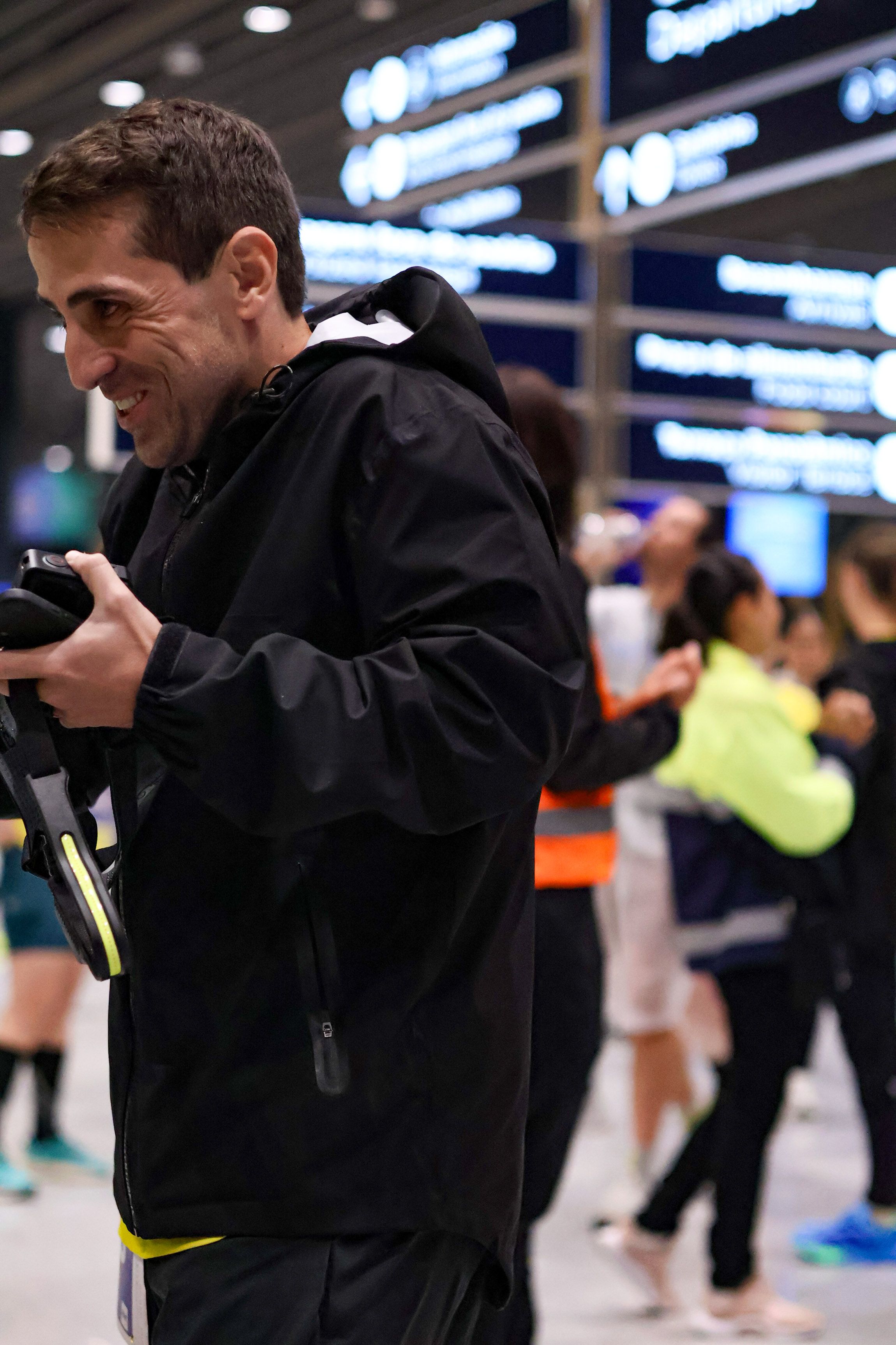 Corredor sorrindo no saguão do aeroporto com o painel de voos ao fundo, vestindo a jaqueta preta impermeável Nord